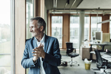 Smiling male entrepreneur with hands clasped looking through window in office - GUSF05301