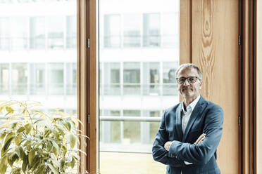 Confident male entrepreneur with arms crossed standing at glass window in office - GUSF05302