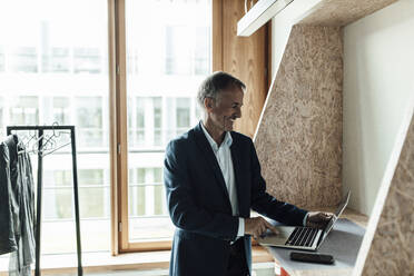 Lächelnder Geschäftsmann, der einen Laptop benutzt, während er in der Lobby eines Büros steht - GUSF05304