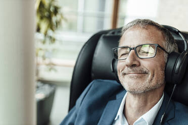 Lächelnder Geschäftsmann mit Brille hört Musik über Kopfhörer, während er auf einem Stuhl im Büroflur sitzt - GUSF05312