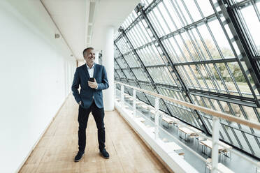 Cheerful businessman with hand in pocket looking away while holding smart phone in corridor - GUSF05317