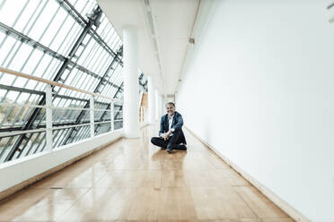 Lächelnder Geschäftsmann auf dem Boden sitzend im Bürokorridor - GUSF05320
