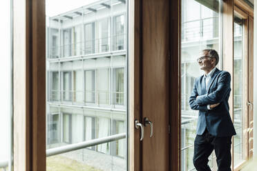 Smiling male entrepreneur with arms crossed looking through glass in corridor - GUSF05323