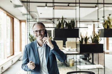 Smiling businessman with eyeglasses talking on telephone while standing in office - GUSF05324
