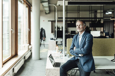 Senior male entrepreneur with arms crossed looking away while sitting by laptop on desk in office - GUSF05328