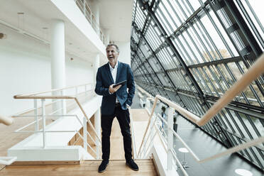 Cheerful businessman holding digital tablet while standing at railing in office corridor - GUSF05335