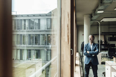 Nachdenklicher Geschäftsmann mit verschränkten Armen, der durch ein Fenster schaut, während er im Büro steht - GUSF05339