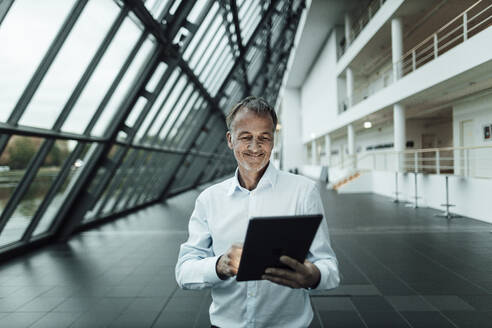 Smiling businessman using digital tablet in office corridor - GUSF05344