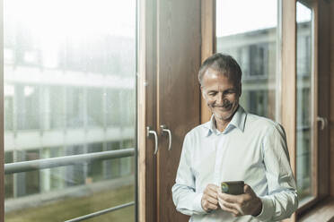 Lächelnder männlicher Unternehmer, der ein Smartphone benutzt, während er am Fenster im Büro steht - GUSF05349