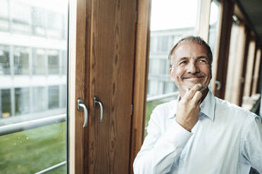 Smiling male entrepreneur with hand on chin looking away while standing at window in office - GUSF05351