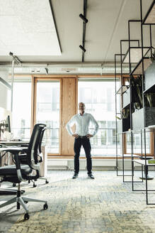 Businessman with hand on hip standing against glass window in office - GUSF05353