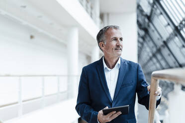 Smiling businessman with digital tablet looking away in office corridor - GUSF05361