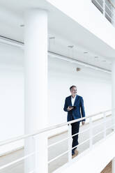 Smiling senior male entrepreneur with digital tablet looking away while walking in office corridor - GUSF05362