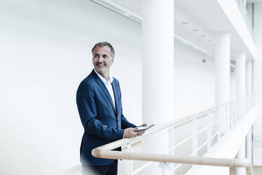 Smiling male business professional holding digital tablet at railing in office corridor - GUSF05365