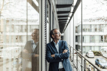 Male entrepreneur with arms crossed looking away while leaning on window in office balcony - GUSF05386