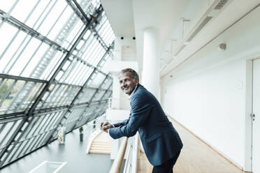 Male business professional smiling while leaning on railing in office corridor - GUSF05388