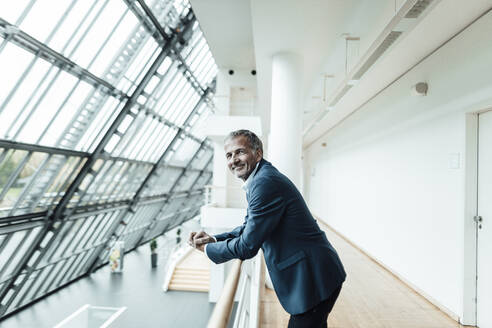 Male business professional smiling while leaning on railing in office corridor - GUSF05388