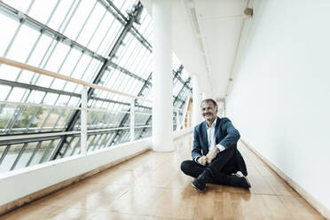 Smiling male business professional sitting on floor in office corridor - GUSF05391