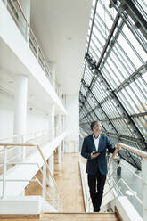 Businessman with digital tablet looking away while moving up from staircase in corridor at office - GUSF05399