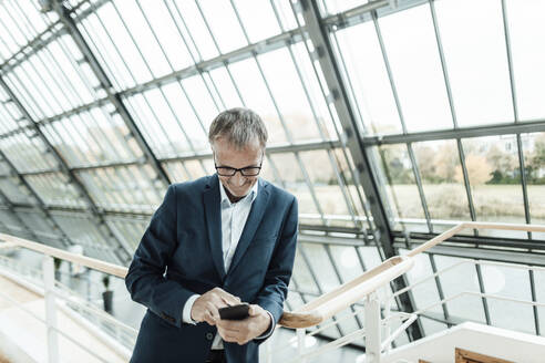 Älterer Geschäftsmann, der sein Smartphone benutzt, während er sich auf das Geländer im Bürokorridor stützt - GUSF05401