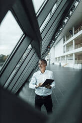 Smiling male entrepreneur holding digital tablet while looking through glass in corridor at office - GUSF05405