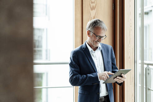 Lächelnder Geschäftsmann, der ein digitales Tablet benutzt, während er vor einem Glasfenster steht - GUSF05409