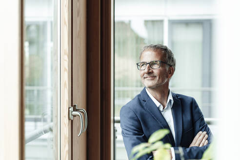 Lächelnder männlicher Geschäftsmann schaut durch ein Fenster im Büro - GUSF05411