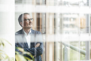 Smiling senior male entrepreneur with arms crossed looking through window in office - GUSF05415