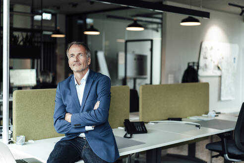 Male business professional with arms crossed sitting on desk in office - GUSF05418