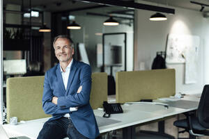 Smiling businessman with arms crossed sitting on desk in office - GUSF05419