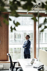 Businessman day dreaming while standing with arms crossed at glass window in office - GUSF05421