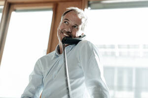 Lächelnder Geschäftsmann, der wegschaut, während er am Telefon gegen ein Fenster im Büro spricht - GUSF05426