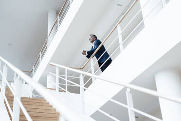 Senior male business professional looking away while leaning on railing in corridor at office - GUSF05461