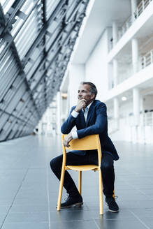 Senior Business Professional mit Hand auf dem Kinn sitzt auf einem Stuhl im Flur im Büro - GUSF05466