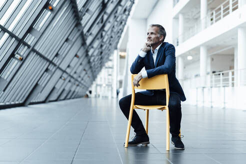 Senior male entrepreneur with hand on chin sitting on chair in office corridor - GUSF05467