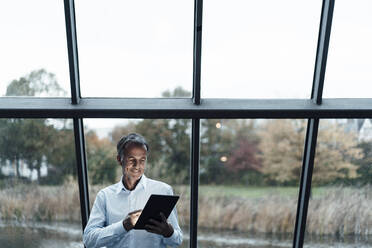 Smiling senior business professional using digital tablet against glass window - GUSF05469
