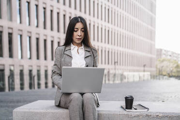 Reife Geschäftsfrau, die an einem Laptop arbeitet, während sie auf einem Stuhl vor einem Gebäude in der Stadt sitzt - JCZF00476