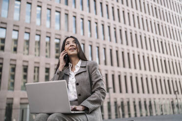 Nachdenkliche Geschäftsfrau, die ein Smartphone benutzt, während sie einen Laptop vor einem Gebäude in der Stadt benutzt - JCZF00477