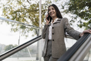 Smiling businesswoman talking on smart phone while standing on escalator in city - JCZF00479