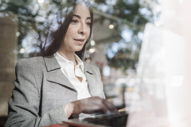 Businesswoman working on laptop while sitting in cafe seen through glass - JCZF00484