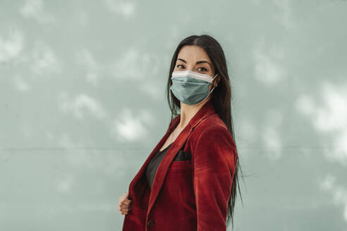 Geschäftsfrau in rotem Blazer mit Gesichtsschutzmaske vor türkisfarbener Wand während einer Pandemie - JCZF00493