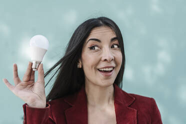 Surprised businesswoman holding light bulb against turquoise wall - JCZF00494
