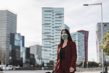 Geschäftsfrau in rotem Blazer mit Gesichtsschutzmaske bei einem Spaziergang in der Stadt während COVID-19 - JCZF00496