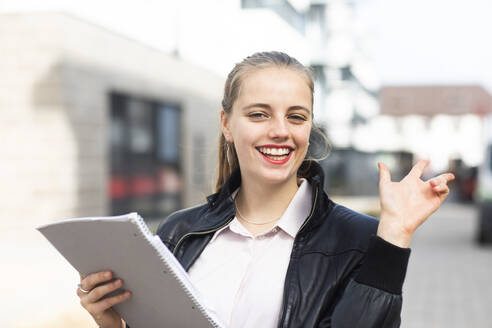 Portrait of young blonde woman standing outdoors with note pad in hand - SGF02779