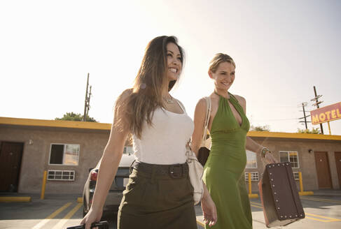 Smiling female friends with luggage walking against motel on sunny day - AJOF01162