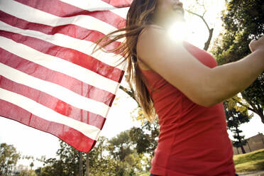 Junge Frau läuft im Park, während sie die amerikanische Flagge an einem sonnigen Tag hält - AJOF01176