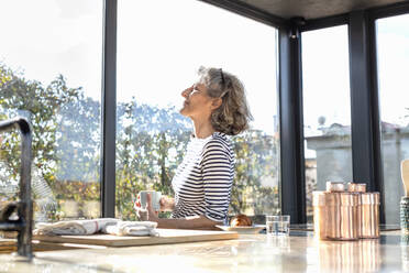 Sonnenlicht fällt auf eine ältere Frau, die mit einer Kaffeetasse in der Küche steht - EIF00415