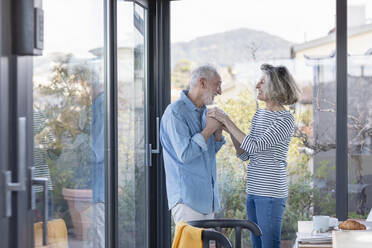 Senior couple holding hands while standing by window at home - EIF00429