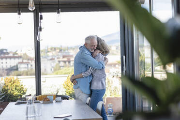 Älteres Ehepaar umarmt sich zu Hause am Fenster stehend - EIF00430