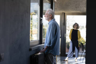 Älterer Mann schaut durch ein Fenster, während er mit einer Frau im Hintergrund zu Hause steht - EIF00431